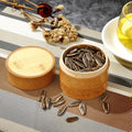 Bamboo food storage box with lid containing sunflower seeds, displayed with dried flowers, oil, and scattered seeds on stripe
