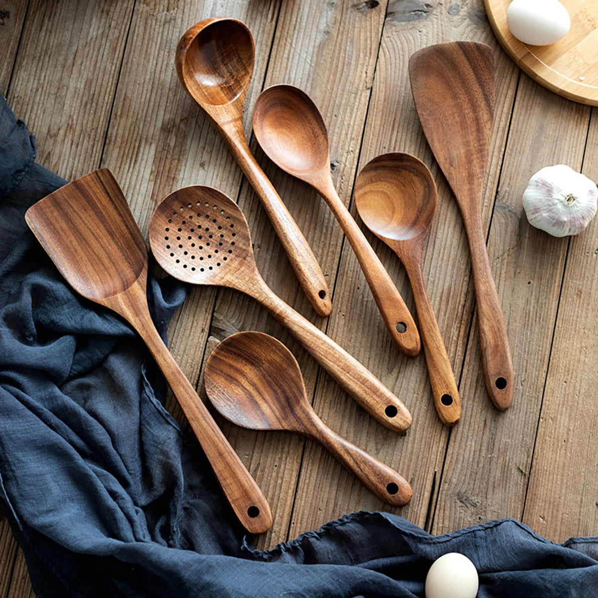 Wooden kitchen utensil set with spatula, spoons, and fork arranged on rustic wood surface with garlic and eggs.