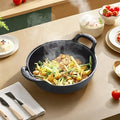 Cast iron double-handle pot with steaming stir-fry dish on beige countertop, surrounded by ingredients and serving bowls.