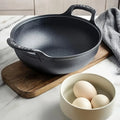 Black cast iron double-handle pot on wooden board with gray linen towel and bowl of eggs on marble countertop.