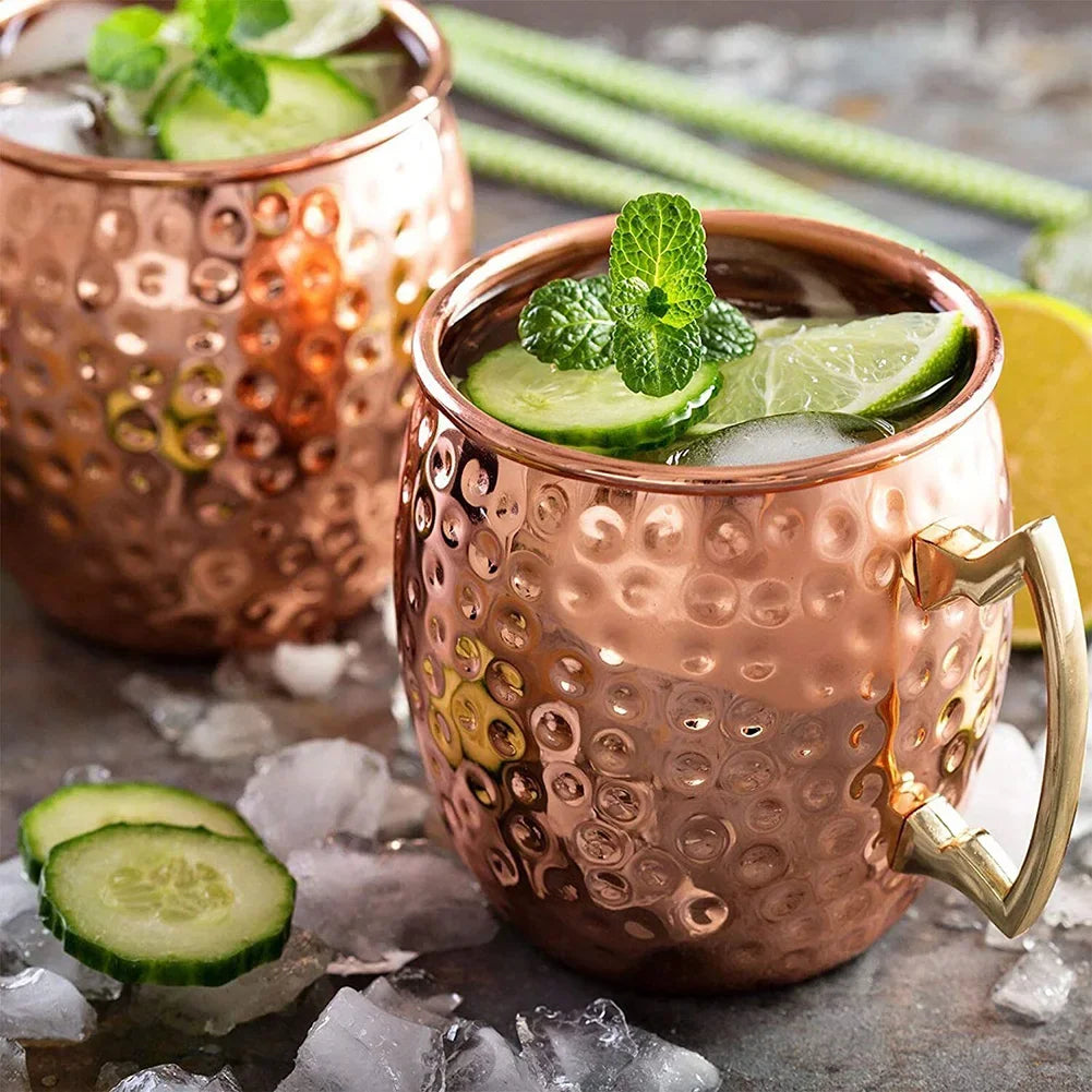 Hammered copper Moscow mule mugs with brass handles, filled with cocktails, lime slices, and fresh mint, set on ice with cucu