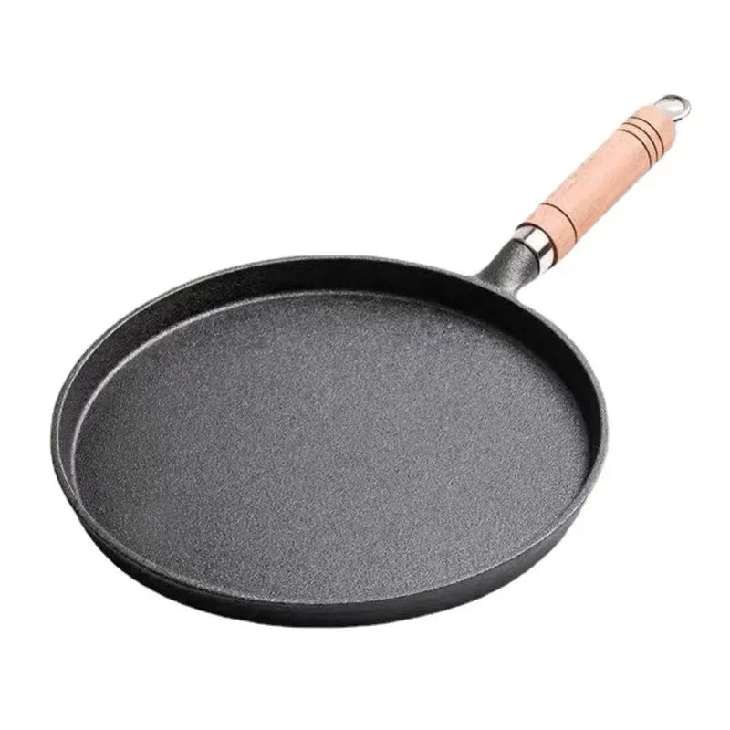 Cast iron skillet with wooden handle cooking crepes on rustic wooden table with fresh berries, juice, and kitchen linens.