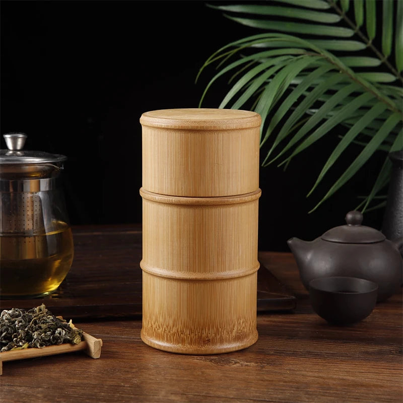 Cylindrical bamboo food storage container with lid and decorative bands, displayed with tea, teapot, and dried herbs on woode