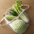 Cotton mesh grocery bag filled with fresh celery, avocado, and zucchini on tan background