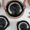 Black earthenware casserole pot with natural woven seagrass handle, lid shown, rustic artisanal design for slow cooking.
