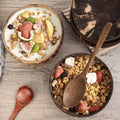 Handcrafted wooden serving bowl filled with granola, fresh fruit, and yogurt on rustic wooden surface with wooden spoon.