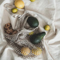Reusable cotton mesh grocery bags filled with fresh produce including avocados, lemons, ginger, and leafy greens on white fab
