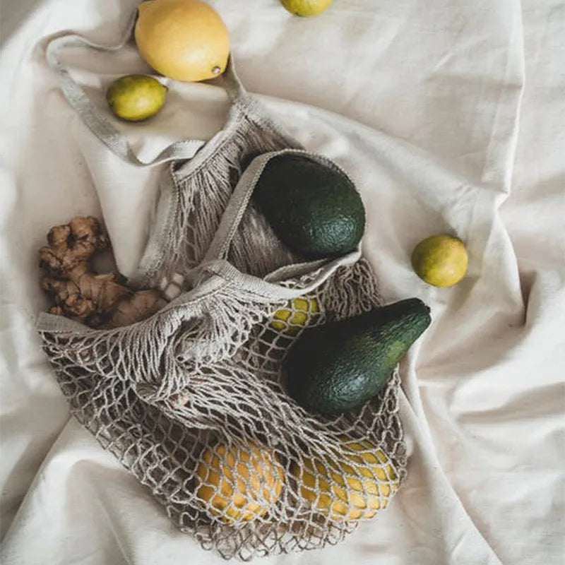 Reusable cotton mesh grocery bags filled with fresh produce including avocados, lemons, ginger, and leafy greens on white fab