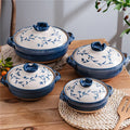 Blue and white ceramic casserole pots with leaf designs, displayed on wooden surface with woven placemats and potted plants.