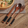 Wooden cutlery set with chopsticks, spoon, and fork featuring black handles, displayed on striped cloth next to wooden bowl o