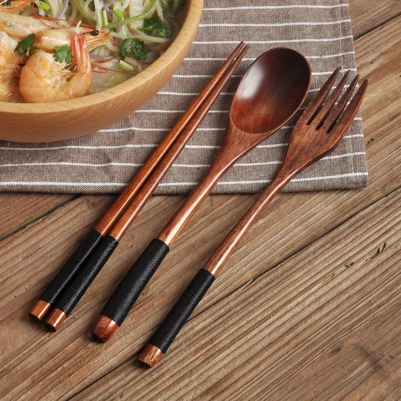 Wooden cutlery set with chopsticks, spoon, and fork featuring black handles, displayed on striped cloth next to wooden bowl o