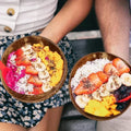 Coconut wood bowls filled with colorful acai bowls, granola, fresh fruit, and coconut flakes, held by two people wearing casu