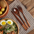 Wooden cutlery set with spoon, fork, and chopsticks on striped napkin beside wooden bowl with noodles and boiled eggs.
