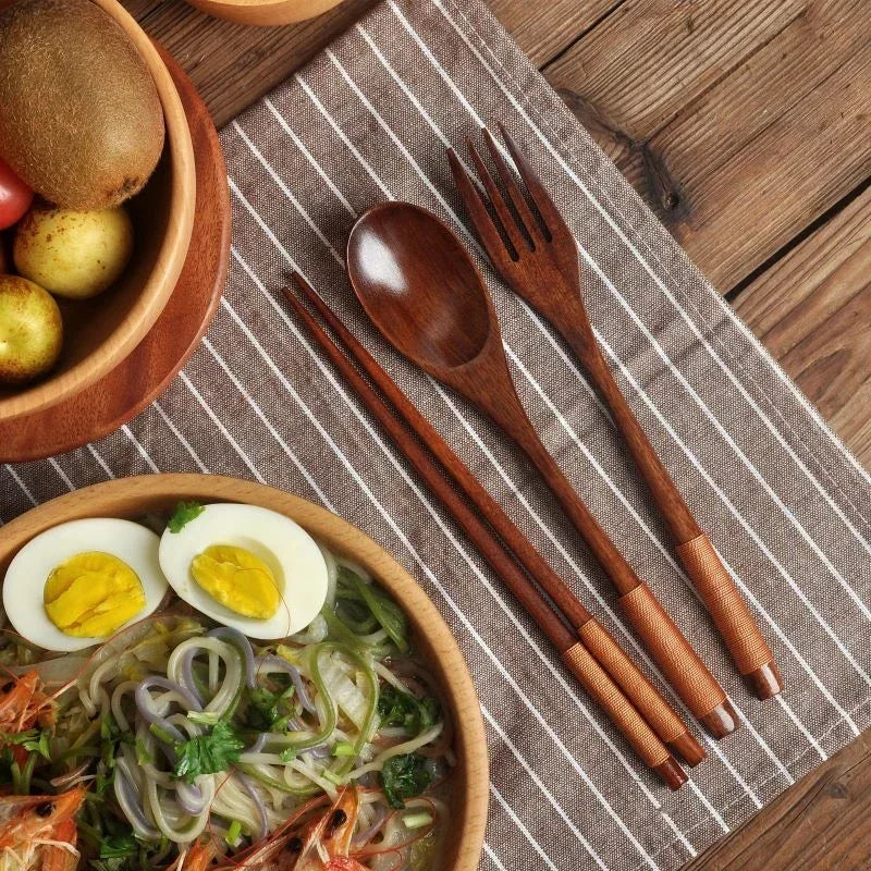 Wooden cutlery set with spoon, fork, and chopsticks on striped napkin beside wooden bowl with noodles and boiled eggs.