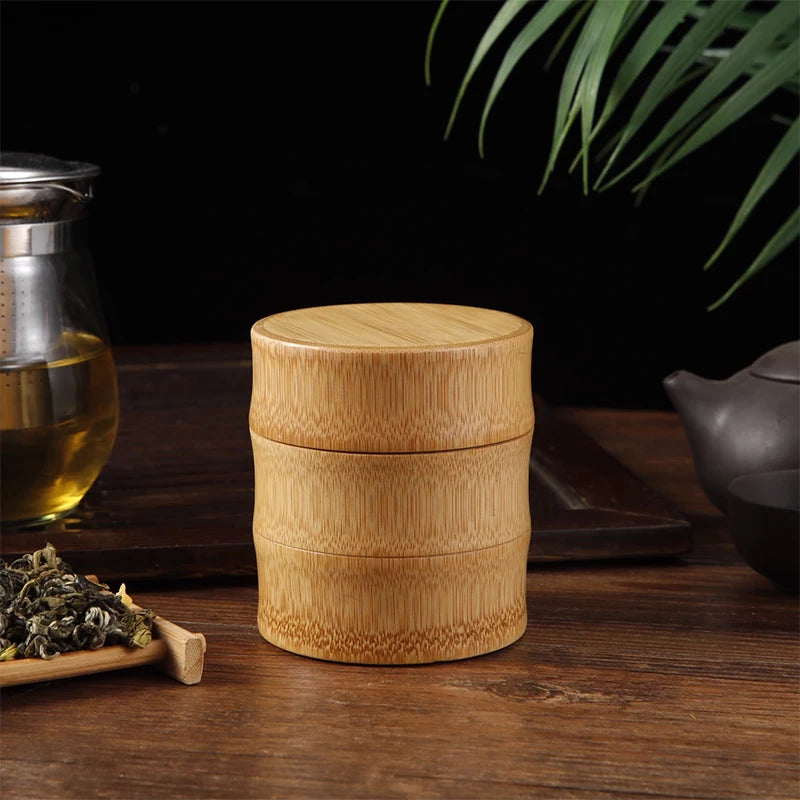 Stacked bamboo food storage container with natural finish, displayed on wooden surface with tea leaves, glass jar, and potted
