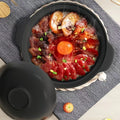 Black ceramic clay cooking pot with lid, filled with raw beef slices, mushrooms, carrot, and green onions on textile surface.