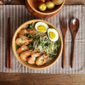 Wooden cutlery set with chopsticks and spoon displayed beside an Asian noodle bowl with shrimp, eggs, and greens on striped p