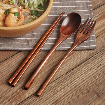 Wooden cutlery set with chopsticks, spoon, and fork on striped linen napkin beside Asian noodle bowl on wooden table.