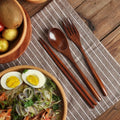 Wooden cutlery set with spoon, fork, and chopsticks on striped napkin, displayed with wooden bowl of noodles and eggs on rust