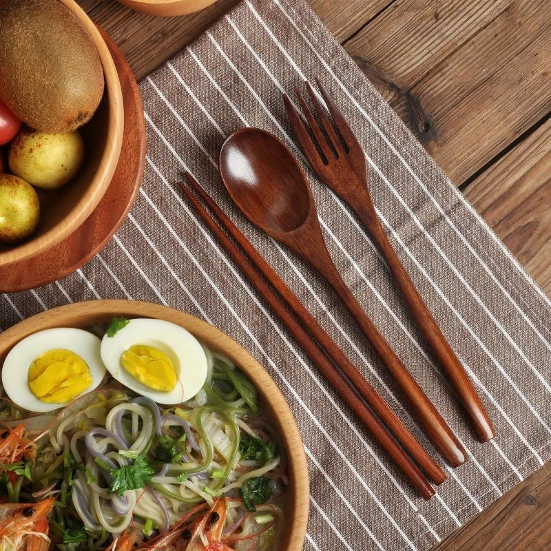 Wooden cutlery set with spoon, fork, and chopsticks on striped napkin, displayed with wooden bowl of noodles and eggs on rust