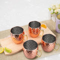 Four hammered copper Moscow Mule mugs with black interior linings and brass handles on a wooden board with lime slices.