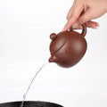 Brown clay teapot being held and pouring water into a black bowl, showing handmade ceramic craftsmanship and spout design.
