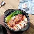Black ceramic clay cooking pot filled with rice, sliced meat, and bok choy, displayed with garlic and blue cloth on wooden su