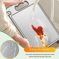 Stainless steel cutting board being rinsed under water, showing easy-clean mesh surface with red sauce residue and slip-resis