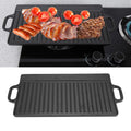 Cast iron dual-sided griddle with grilled meats and tomatoes, showing ridged cooking surface and flat side with handles.