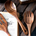 Three wooden kitchen utensils (spoon, spatula, fork) displayed on dark wood surface with vintage book and woven basket.