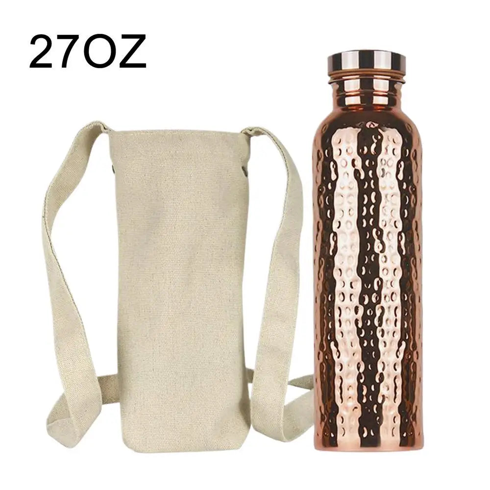 Copper water bottle with hammered design next to cream canvas carry bag with shoulder straps, 27oz capacity