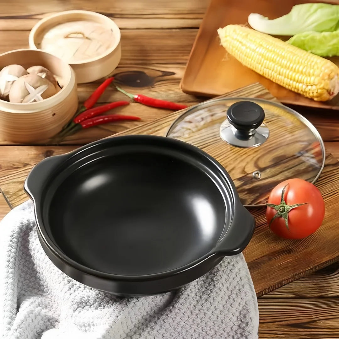 Black ceramic clay cooking pot with glass lid on wooden surface, surrounded by fresh vegetables and cooking ingredients.