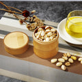 Bamboo food storage container filled with pistachios, displayed with nuts, olive oil, dried flowers, and textured fabric on g