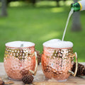 Copper Moscow mule mugs with hammered finish and brass handles, topped with foam, one being filled from green bottle, outdoor