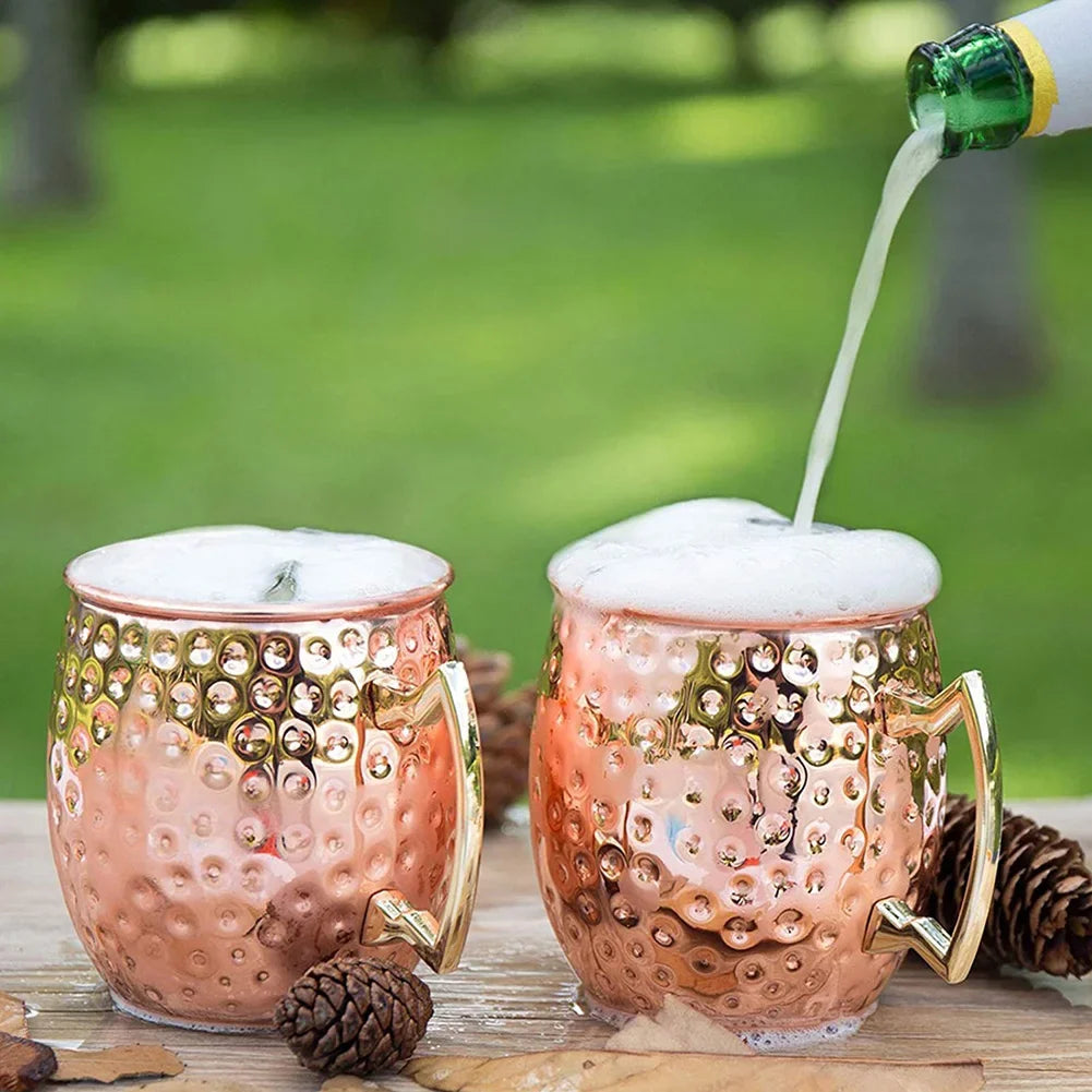 Copper Moscow mule mugs with hammered finish and brass handles, topped with foam, one being filled from green bottle, outdoor