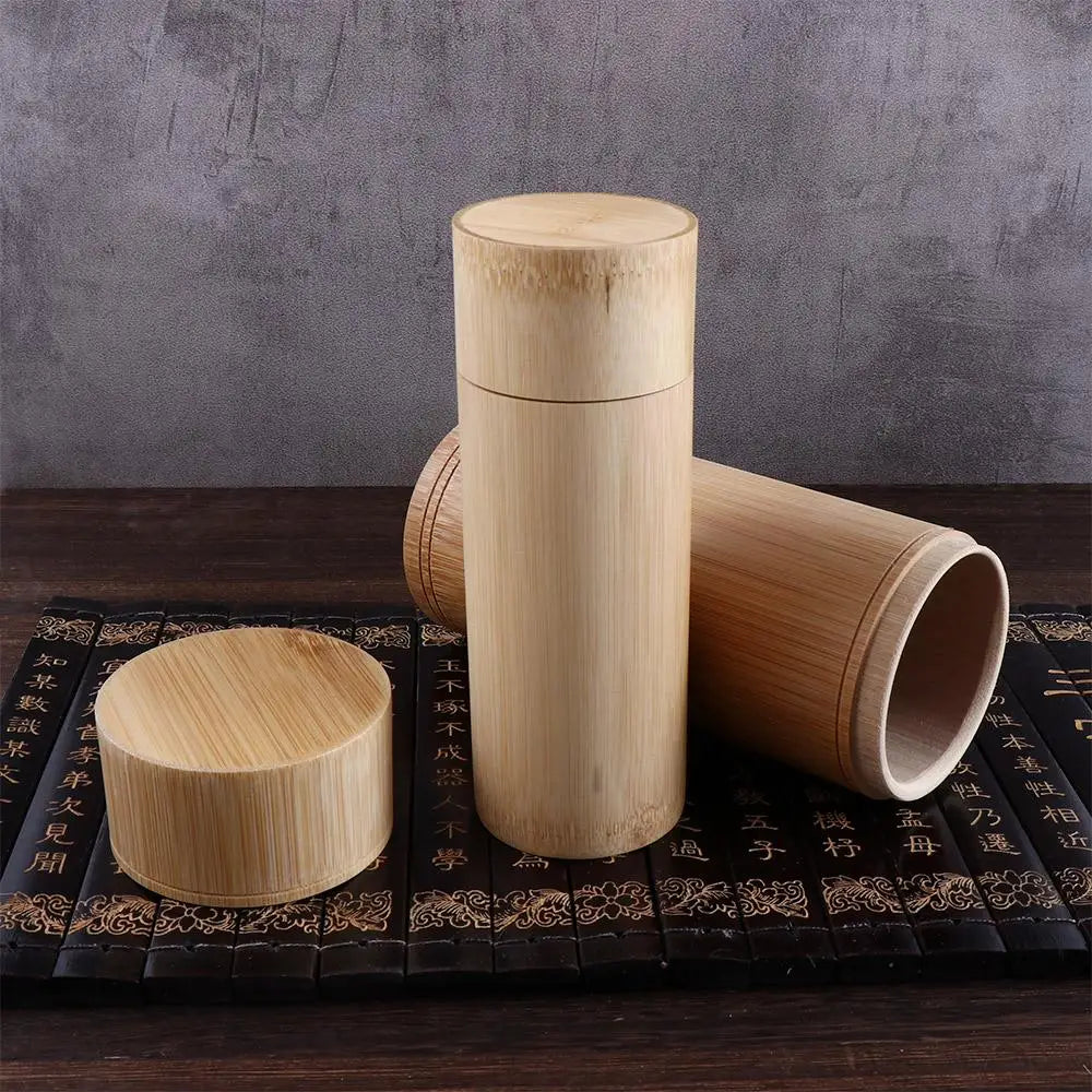 Three handmade bamboo tea storage canisters with wooden lids on a black decorative mat, displayed against a gray textured bac