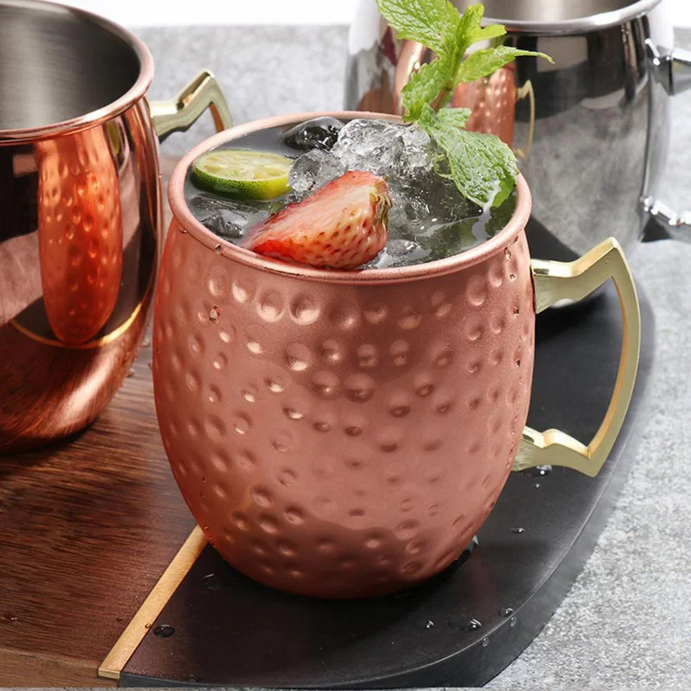 Copper Moscow mule mug with hammered finish, brass handle, filled with ice, strawberry, lime, and mint garnish on black slate