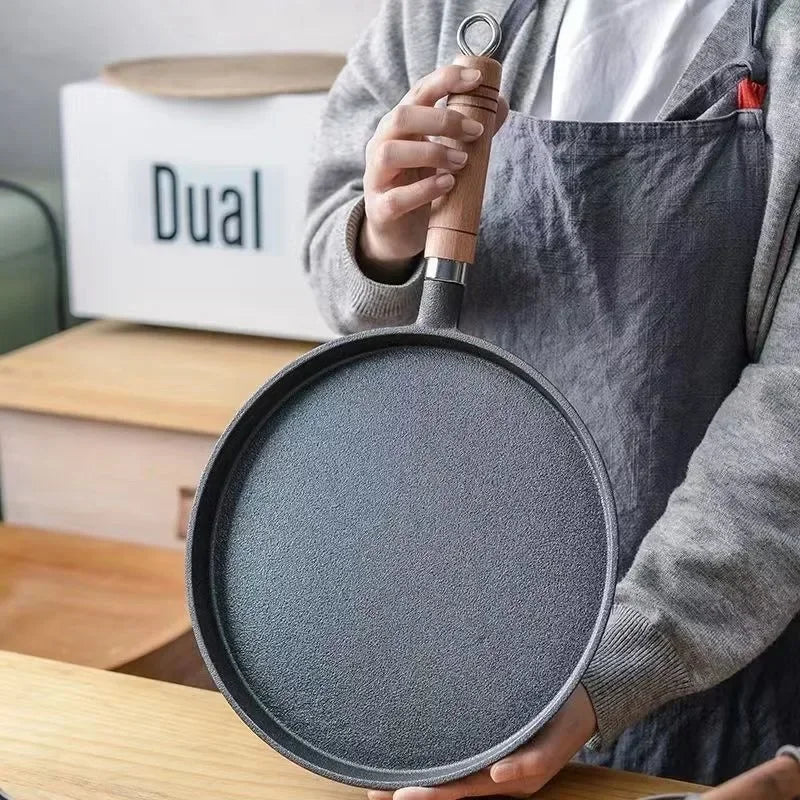 Cast iron egg skillet with dark gray finish, held by hands wearing apron, displayed with Dual brand packaging in background