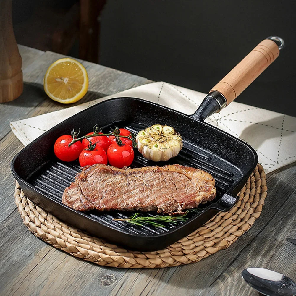 Cast iron square grill pan with wooden handle cooking steak, tomatoes, and garlic on woven trivet, lemon slice nearby.