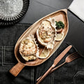 Palm leaf-shaped wooden serving tray with two oysters, lemon slices, and fresh herbs, displayed with copper utensils on dark 
