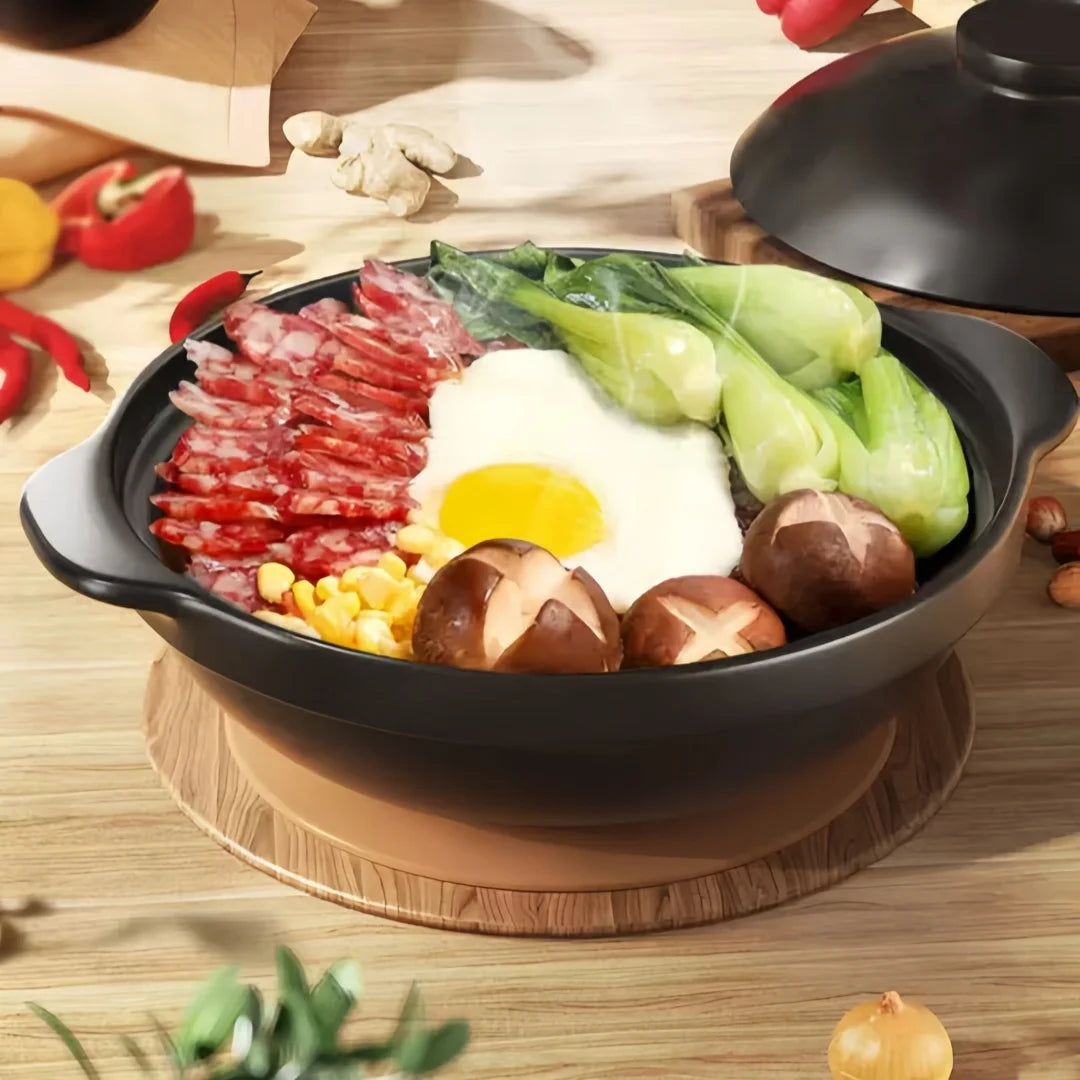 Black ceramic clay cooking pot with wooden base, filled with sliced meat, egg, vegetables and corn on wooden table.