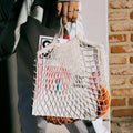 Cotton mesh grocery bag holding fresh oranges and vegetables, displayed against brick wall background