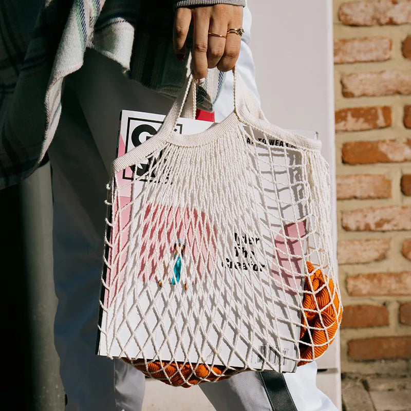 Cotton mesh grocery bag holding fresh oranges and vegetables, displayed against brick wall background