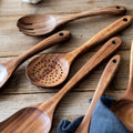 Wooden kitchen utensils set including spatula, slotted spoon, and fork displayed on wooden surface with natural grain detail.
