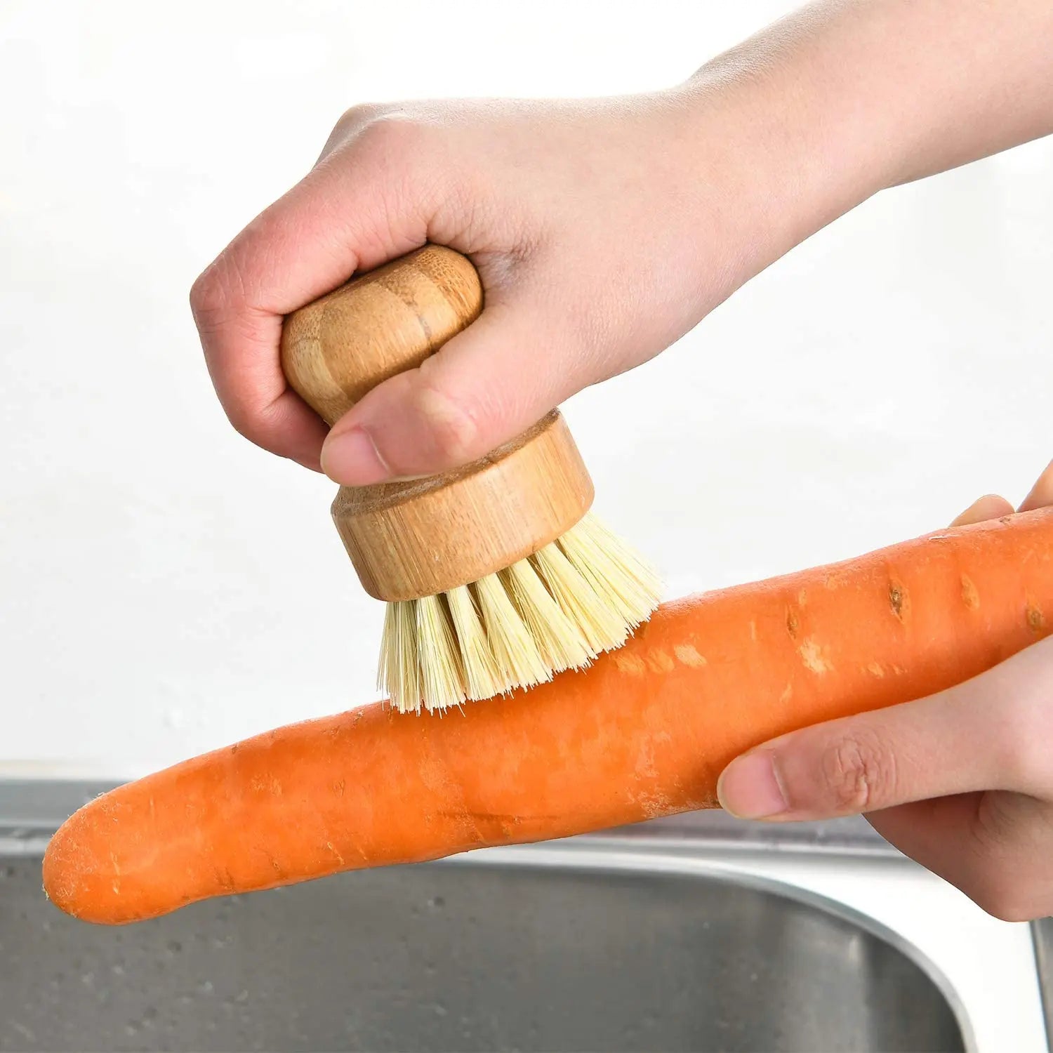 Bamboo dish brush with natural bristles scrubbing a fresh carrot over a gray bowl, demonstrating eco-friendly kitchen cleanin