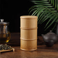 Bamboo food storage box with lid on wooden table, surrounded by tea infuser, black teapot, and green plant leaves.