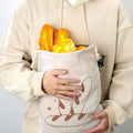 Person holding cream linen bread bag with bread loaf illustration, filled with golden baguettes and rolls
