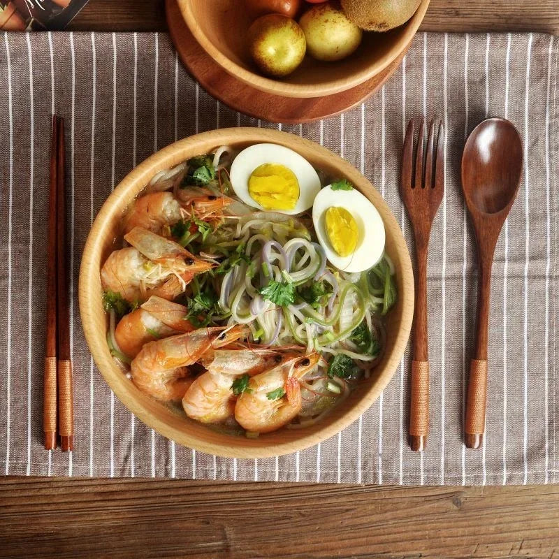 Wooden cutlery set with chopsticks and spoon displayed beside a ramen bowl with noodles, shrimp, and egg on striped placemat.