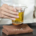 Hand holding glass pitcher of oil on organic cotton cleaning cloth, demonstrating absorbency and eco-friendly kitchen use.