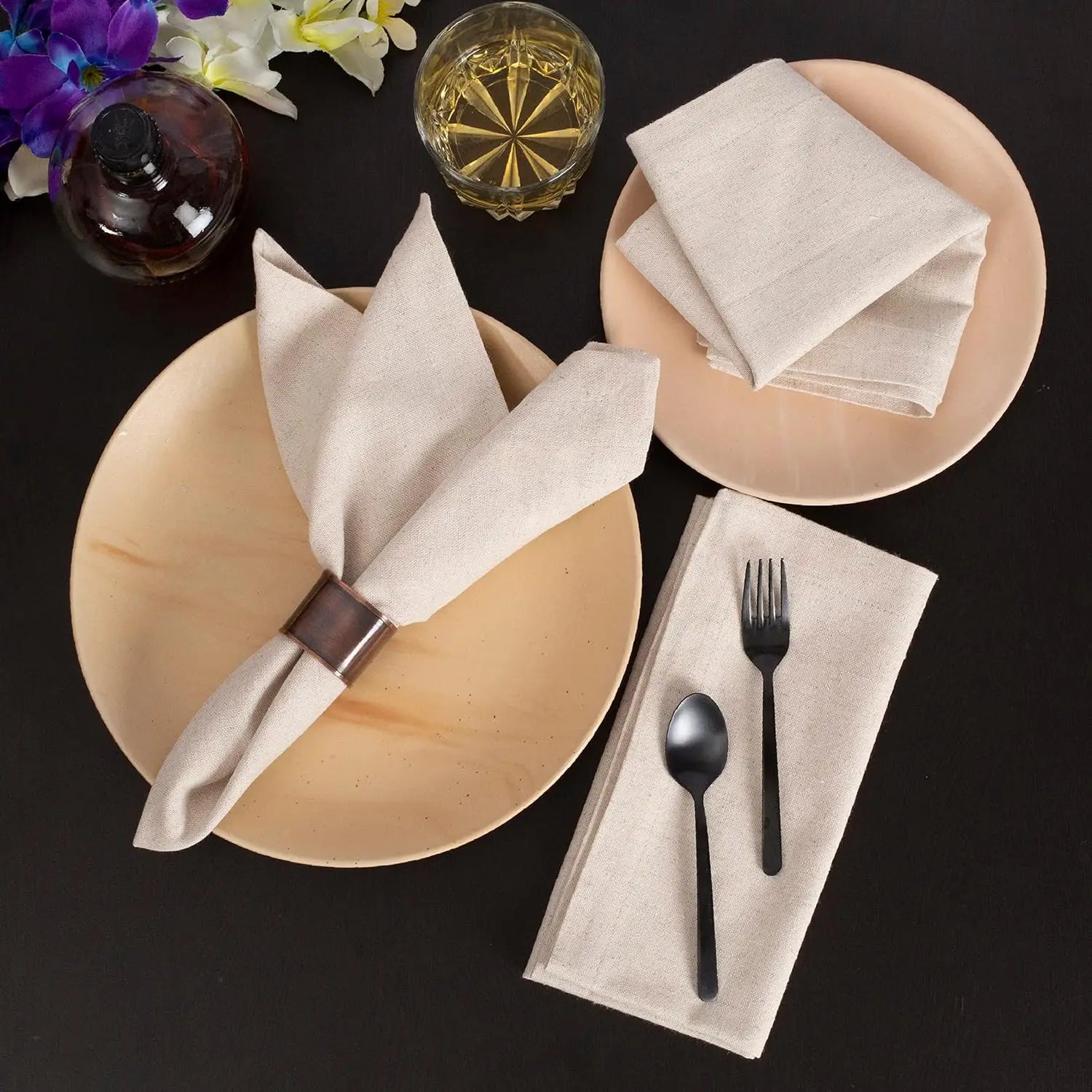 Beige cotton linen napkins styled on plates with black utensils, flowers, and glassware on dark background